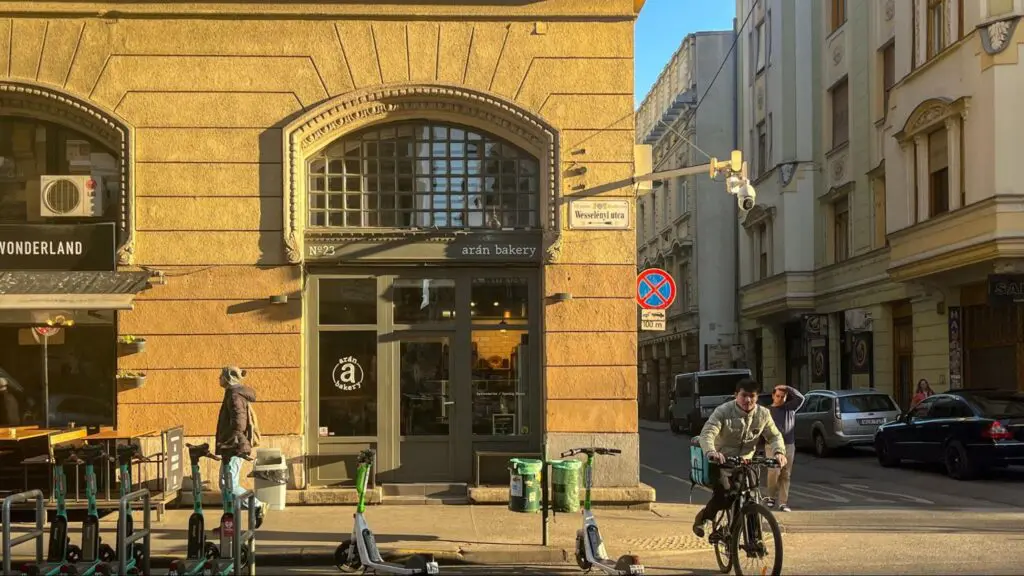 Street view of Arán Bakery located on Wesselényi utca in Budapest, with a warm-toned historic facade, arched windows, and scooters parked out front. Pedestrians and a cyclist pass by in the late afternoon sunlight