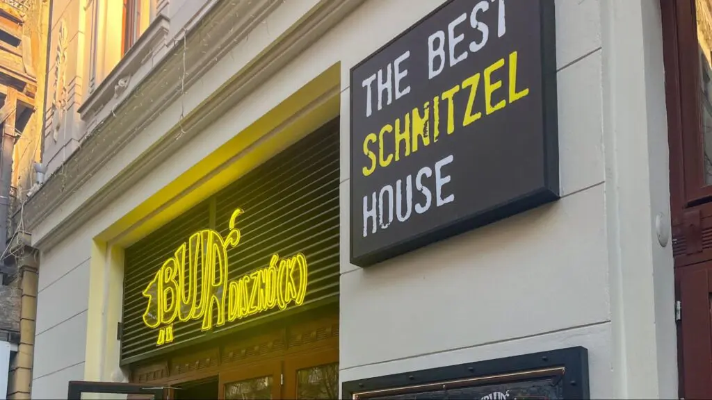 Street view of the entrance to BUNSZNÓ(K), a restaurant with a bright yellow neon sign and a bold black-and-white sign reading 'The Best Schnitzel House' mounted on a white building facade