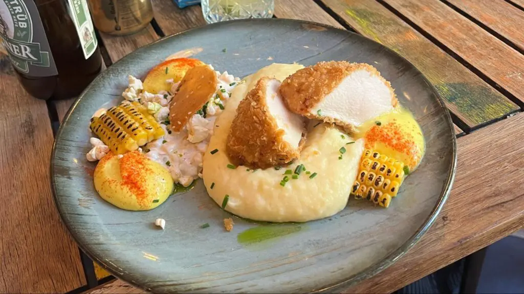 Gourmet dish featuring sliced breaded chicken breast served on creamy mashed potatoes, with grilled corn, dollops of yellow sauce sprinkled with paprika, popcorn, and garnished with chopped chives on a ceramic plate.