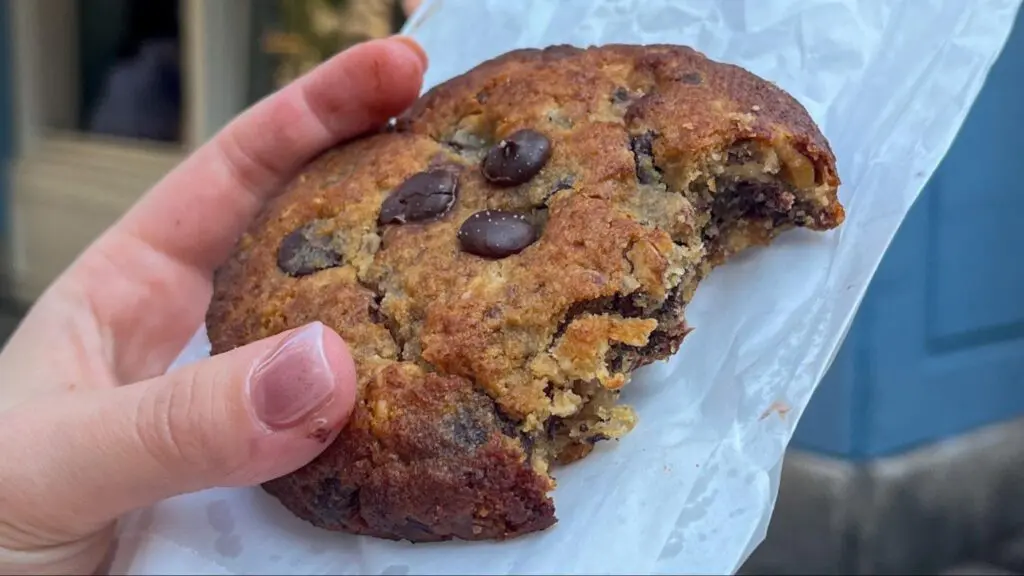 Close-up of a hand holding a partially eaten chocolate chip cookie with a golden-brown crust and melted chocolate chunks, resting on a piece of white parchment paper