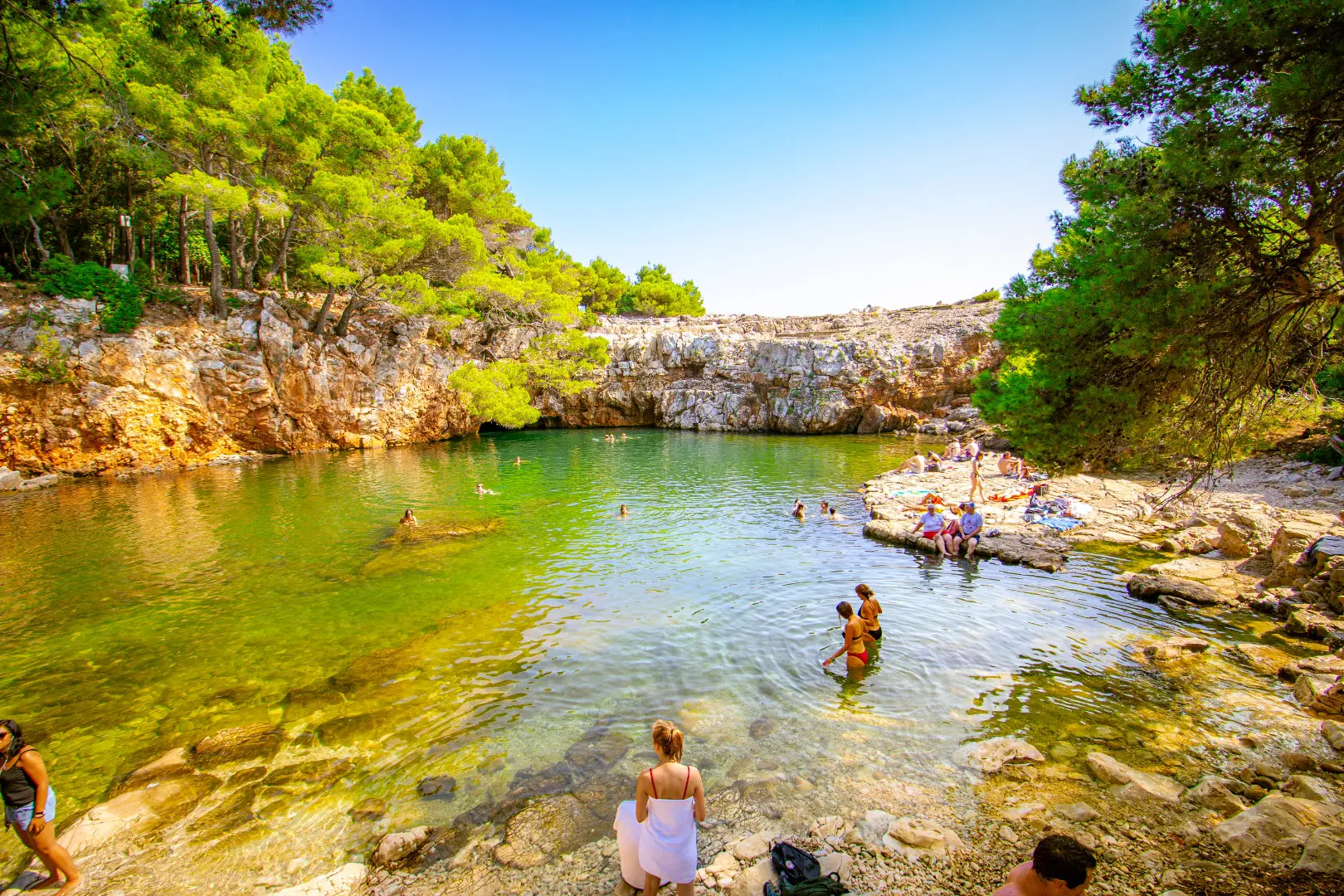 People swimming and relaxing around the vibrant green waters of the Dead Sea (Mrtvo More) lagoon on Lokrum Island, surrounded by rocky cliffs and pine trees.
