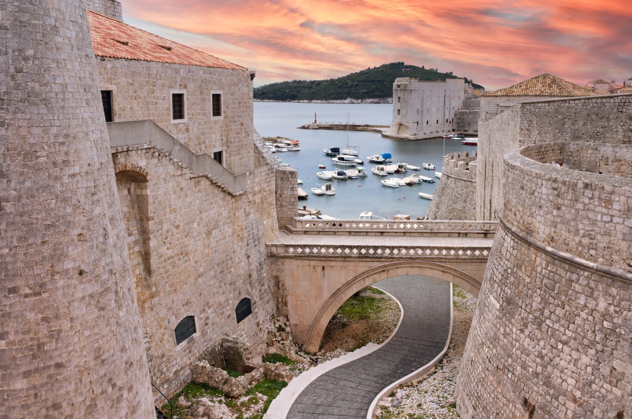 Historic stone walls and a bridge in Dubrovnik, Croatia, with a scenic harbor full of boats and a colorful sunset sky.
