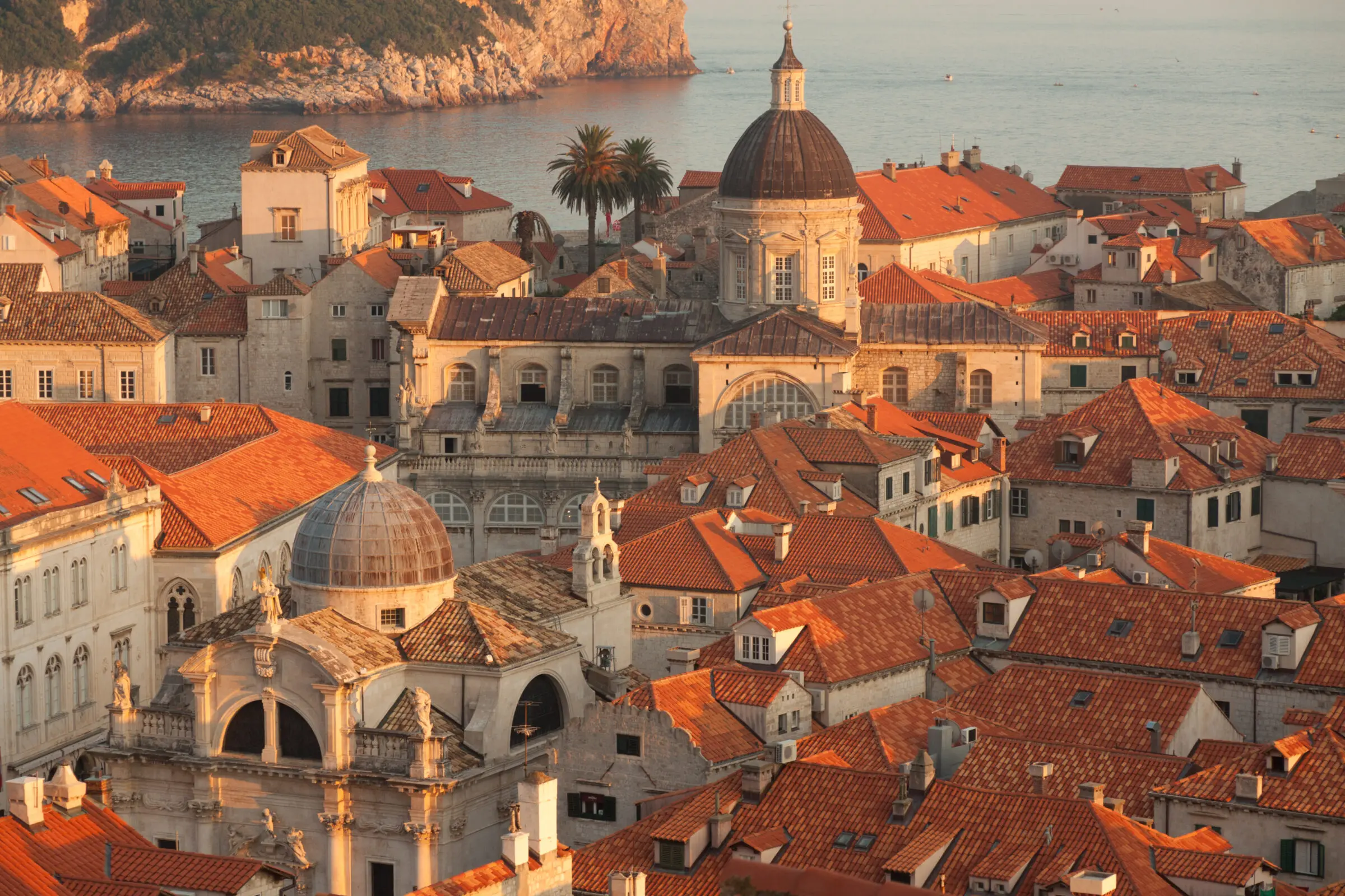 A panoramic view of Dubrovnik’s Old Town in Croatia at sunset, featuring a cluster of historic stone buildings with orange terracotta roofs, prominent church domes, and the calm Adriatic Sea in the background.