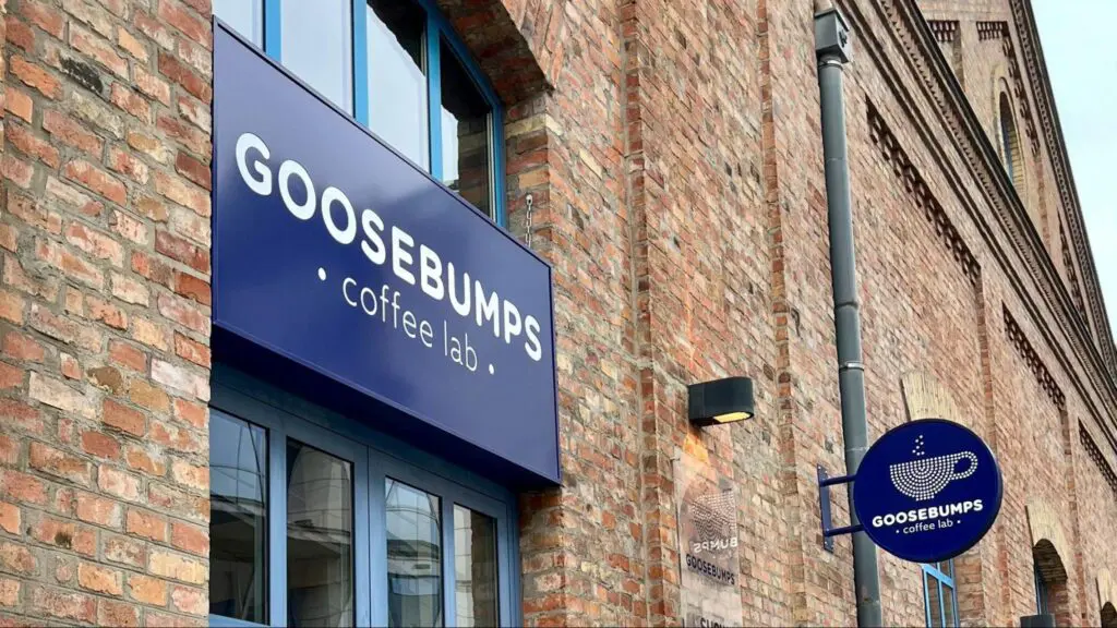 Exterior of Goosebumps Coffee Lab, a specialty coffee shop with a modern blue sign on a historic brick building. The logo features a minimalist coffee cup and steam design."
