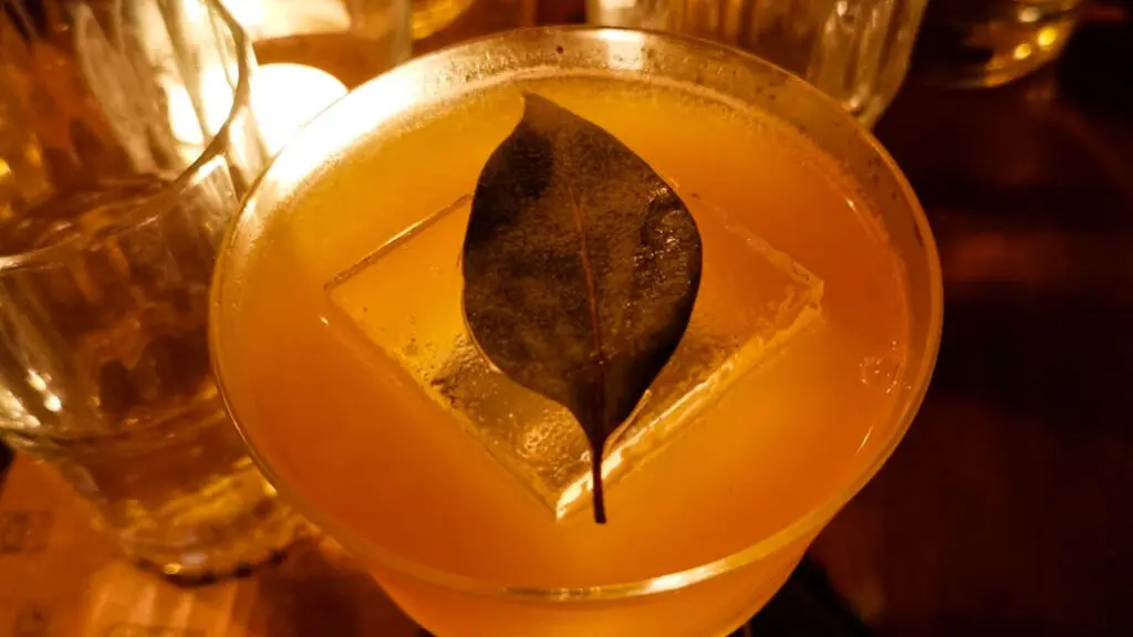 Close-up of a cocktail in a coupe glass, garnished with a single bay leaf resting on a large clear ice cube, with warm ambient lighting and glassware in the background