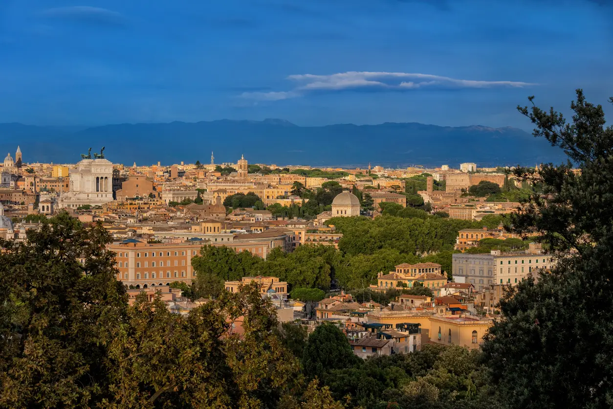 Panoramic view of Rome at sunset with the Altare della Patria, domes of historic churches, and tree-lined rooftops, set against the backdrop of distant mountains