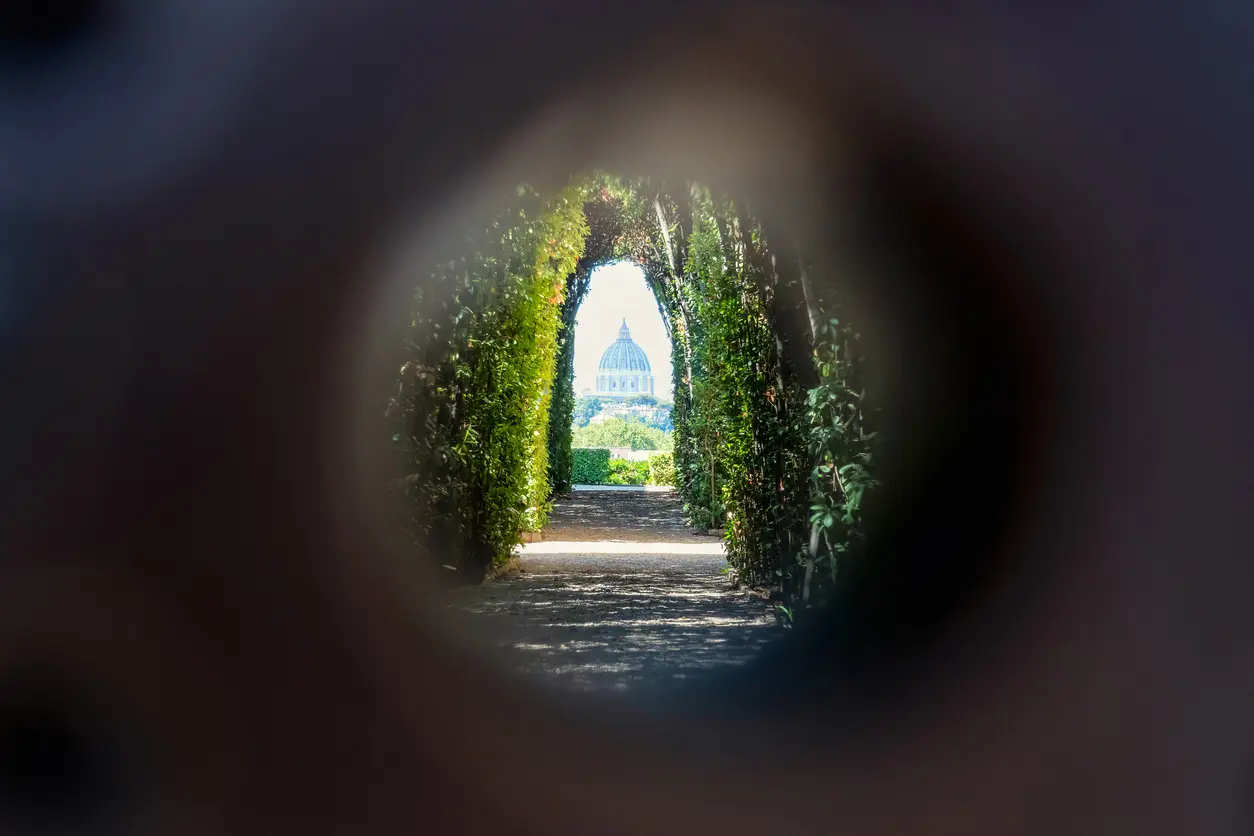 View of St. Peter’s Basilica framed by a keyhole on the Aventine Hill in Rome, with a tree-lined garden path leading to the distant dome