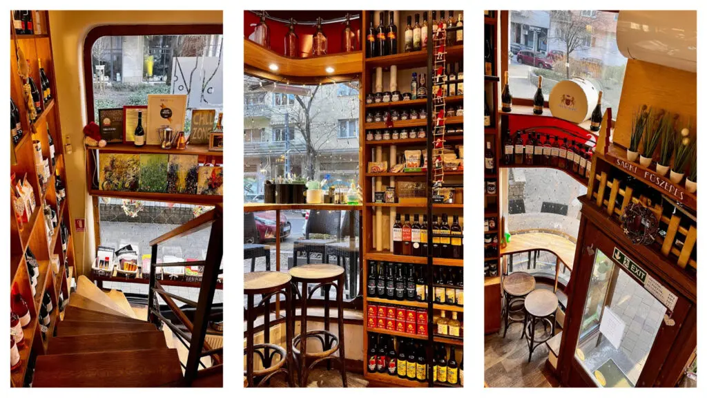 Interior of a cozy European delicatessen or café with wooden stairs, shelves lined with bottles and gourmet products, small round tables with stools near large windows, and warm ambient lighting. The signs and products suggest a Hungarian theme, with the name 'Sarki Fűszeres' visible