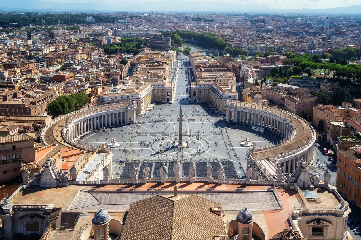 Aerial view of St. Peter’s Square in Vatican City, featuring the central obelisk, colonnades, and the surrounding buildings of Rome, with Castel Sant’Angelo and the Tiber River in the background.