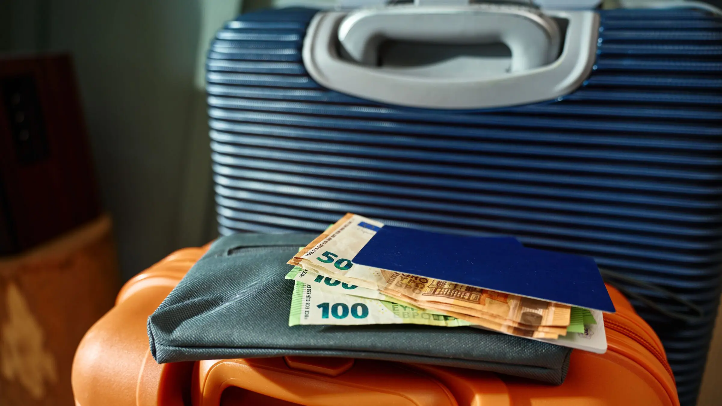 A passport and some Euro banknotes on top of an orange suitcase with a blue suitcase in the background.