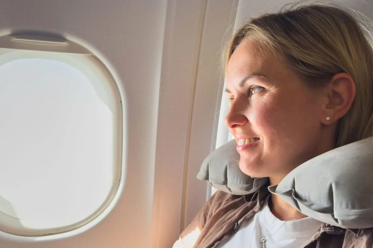 A woman wearing a neck pillow is looking out of an airplane window and smiling.