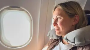 A woman wearing a neck pillow is looking out of an airplane window and smiling.