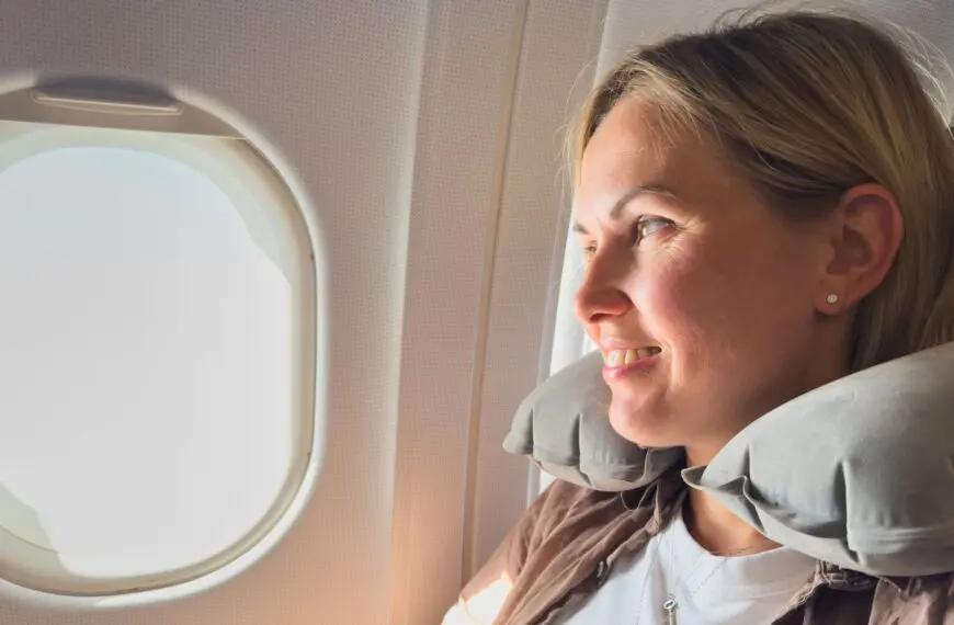 A woman wearing a neck pillow is looking out of an airplane window and smiling.