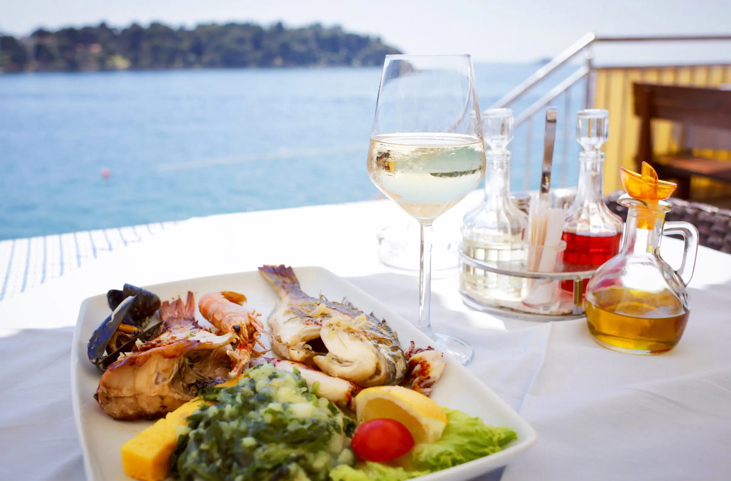 A seafood platter with grilled fish, shrimp, mussels, and vegetables served with a glass of white wine at an outdoor seaside restaurant.