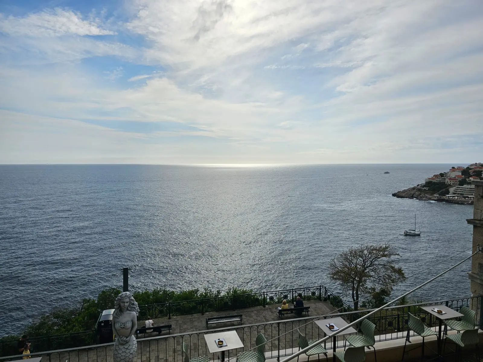 A serene coastal view from a terrace overlooking the Adriatic Sea, with a few diners seated, a mermaid statue in the foreground, and a sailboat near the rocky shoreline of Dubrovnik.
