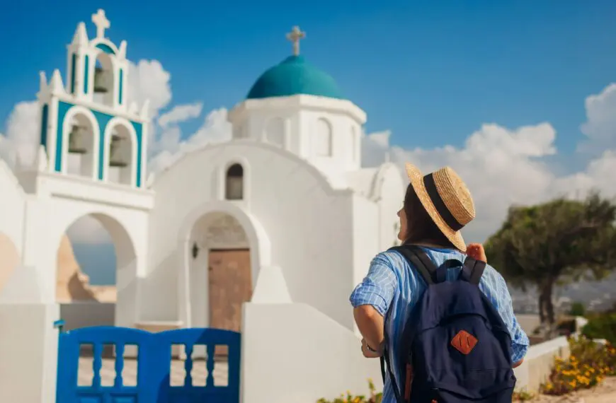 A woman with her back turned to the camera is admiring a whitewashed church in Santorini.