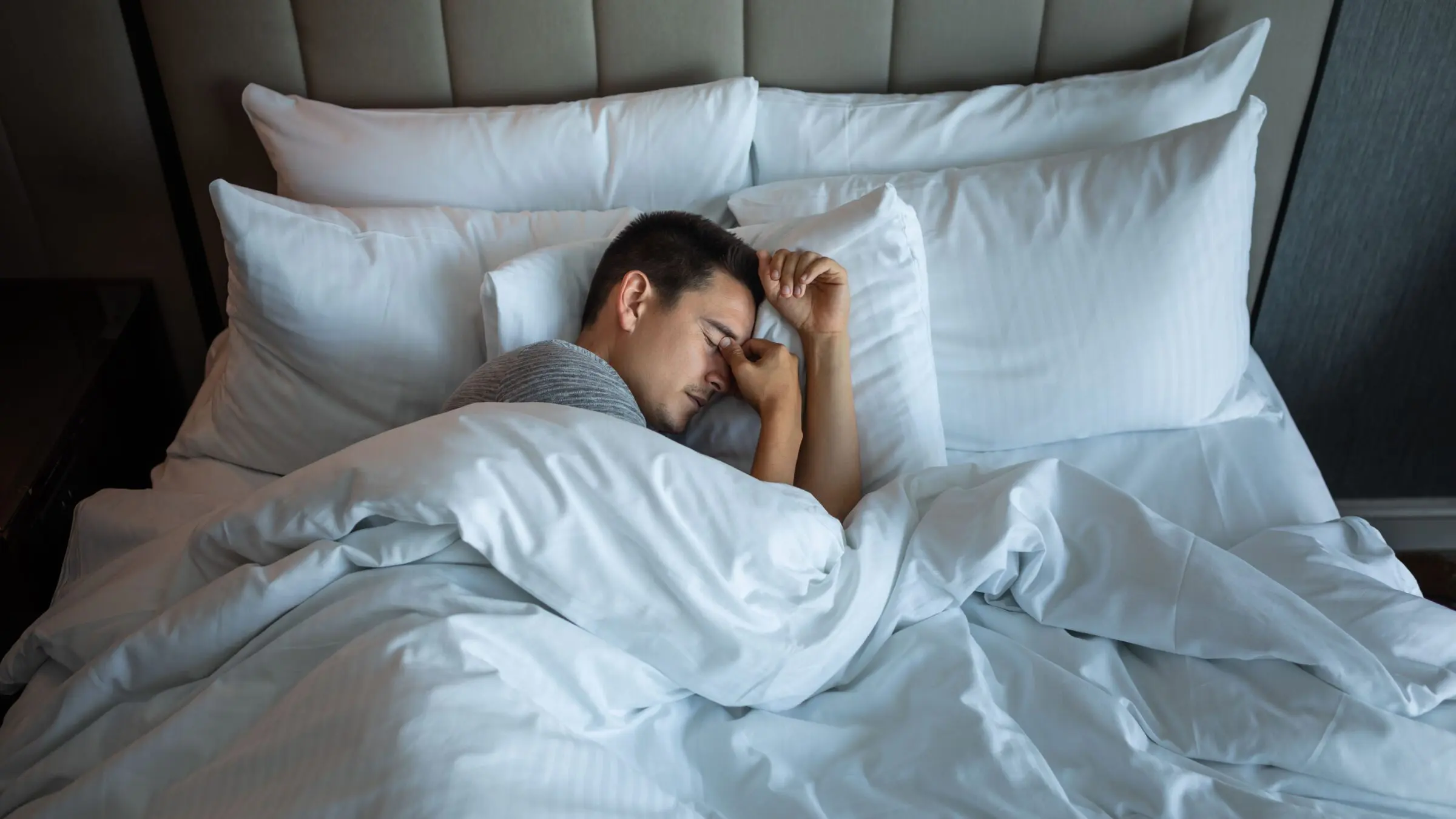A man trying to sleep in a bed with white sheets and white pillows.