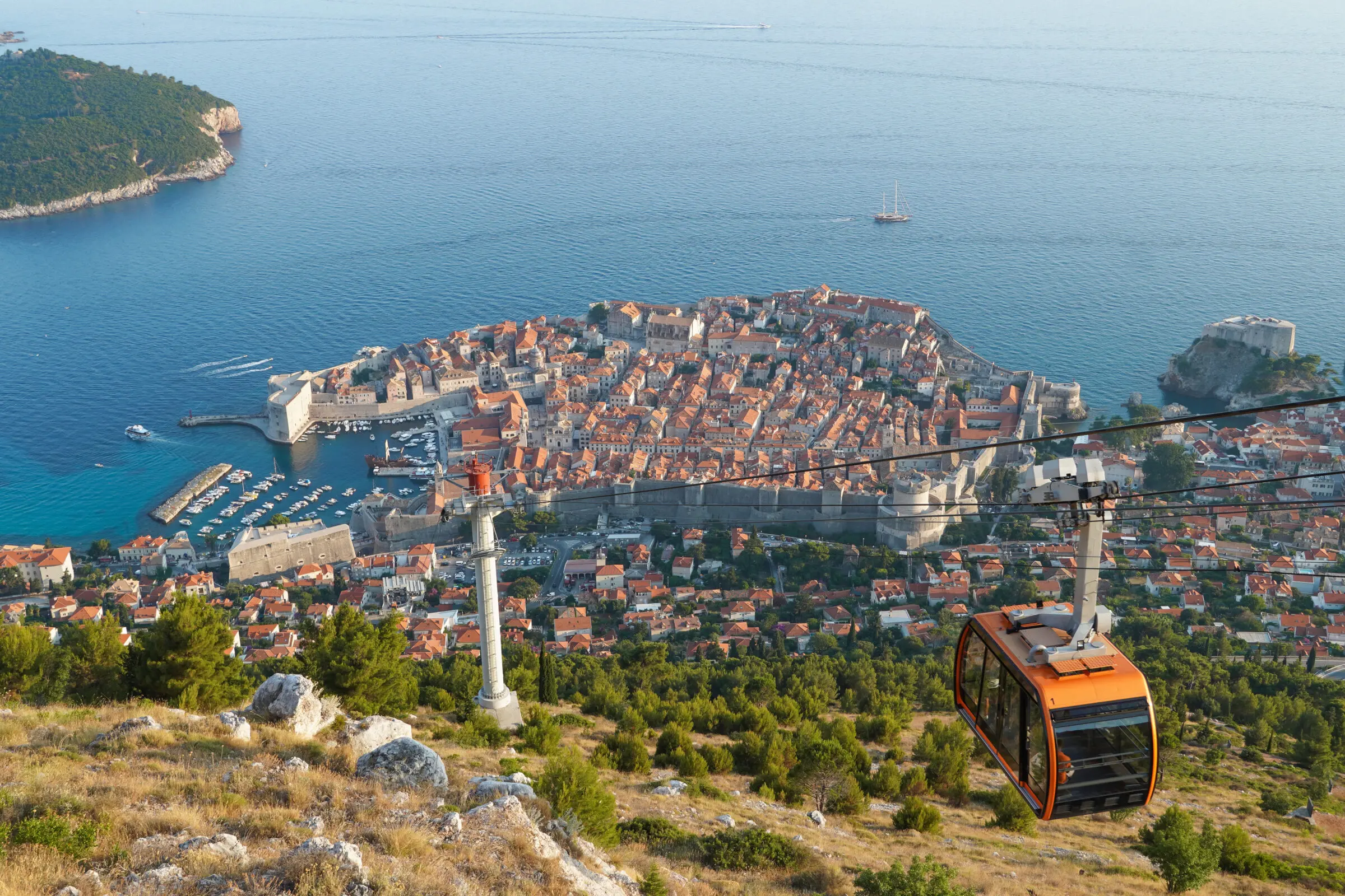 A panoramic view of Dubrovnik’s Old Town and coastline from Mount Srđ, with a bright orange cable car descending toward the city.