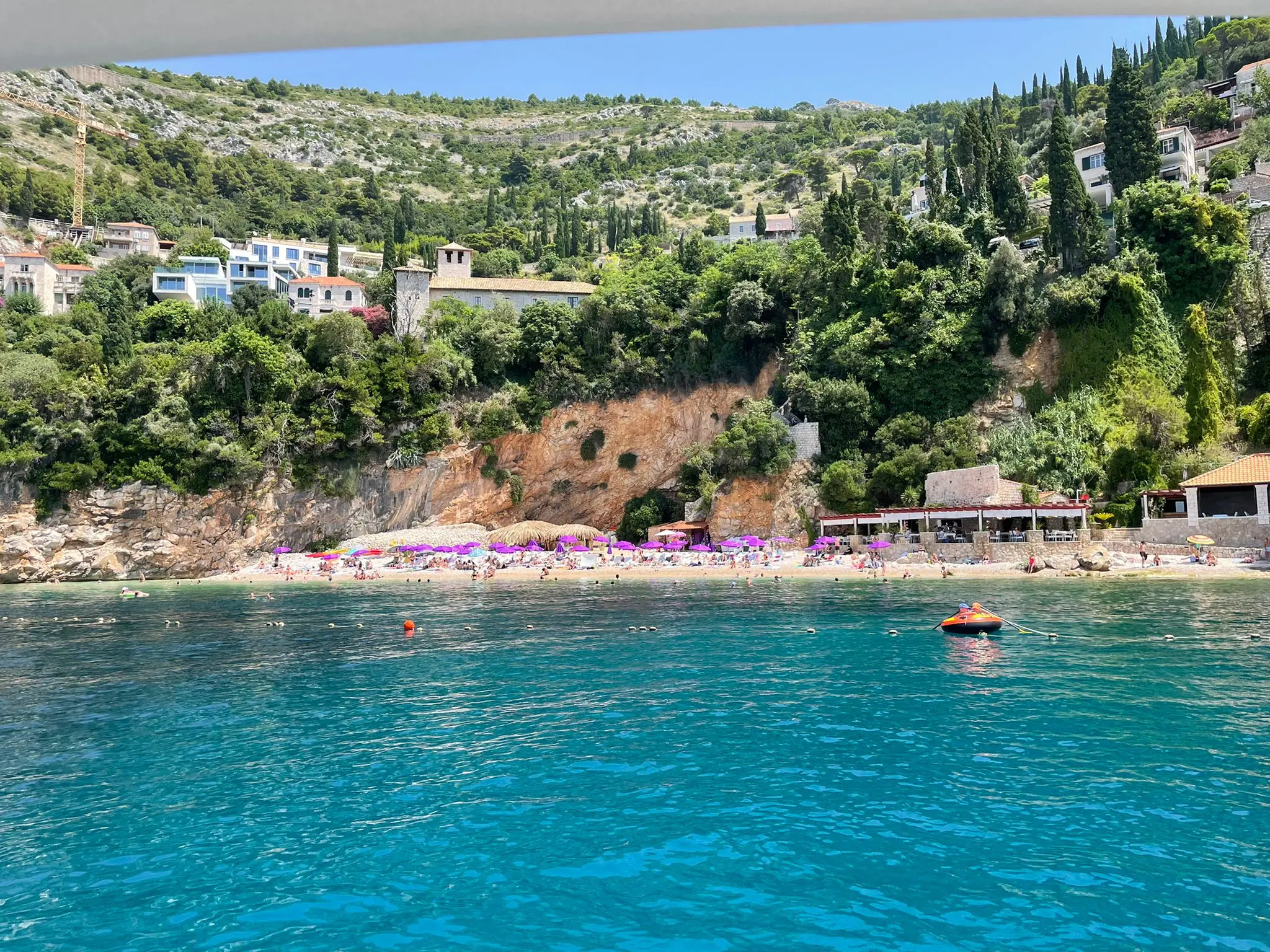 A picturesque beach near Dubrovnik with clear turquoise water, a rocky cliff backdrop, and purple umbrellas lining the sandy shore.
