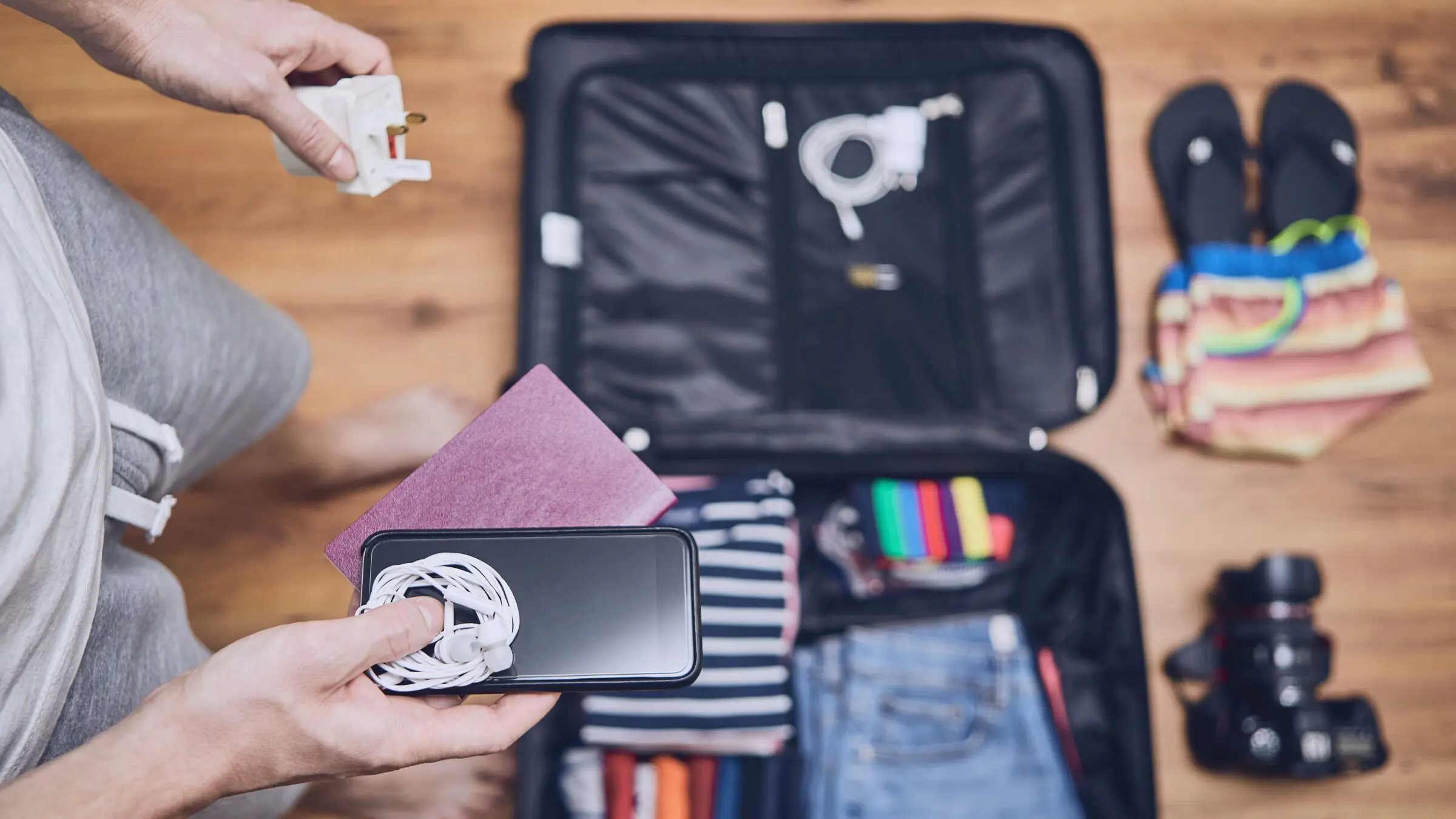 A young man packing clothing, flip flops, a camera and other things in a suitcase on a hardwood floor.