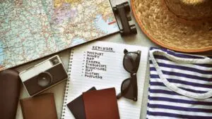 An open notebook with a travel checklist next to a map, a camera, sunglasses, a straw hat, binoculars and a folded striped T-Shirt.