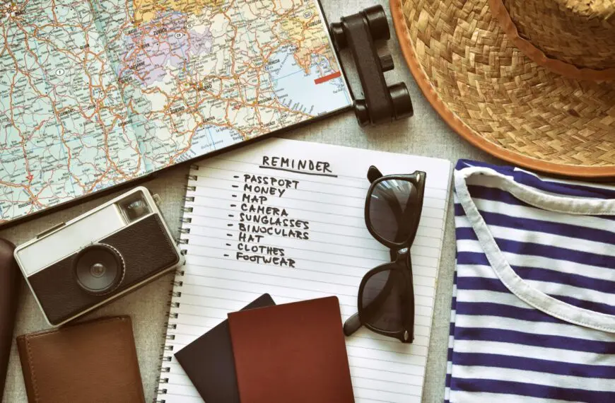 An open notebook with a travel checklist next to a map, a camera, sunglasses, a straw hat, binoculars and a folded striped T-Shirt.
