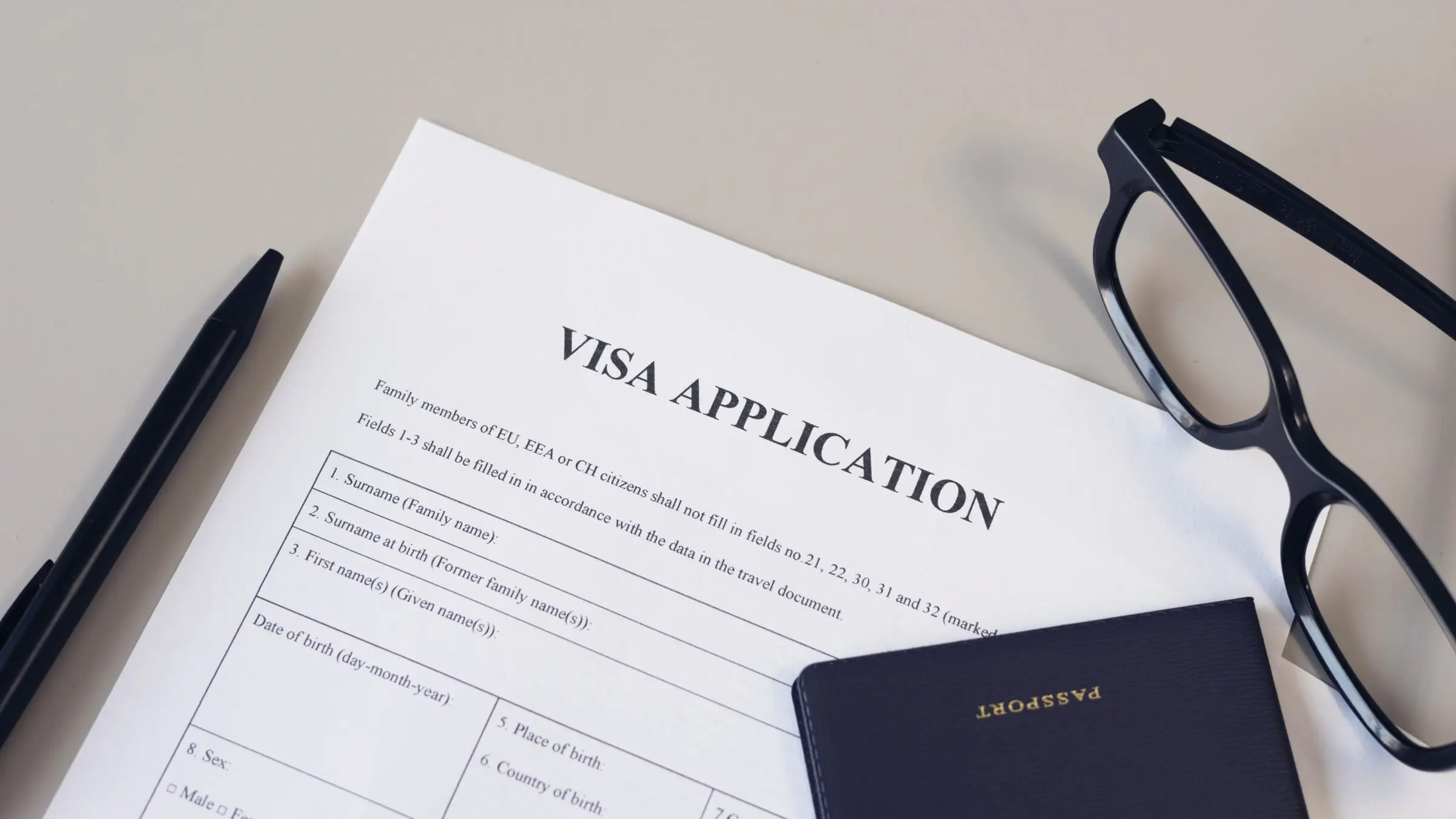 A visa application form, a pen, reading glasses and a passport on a white desk.