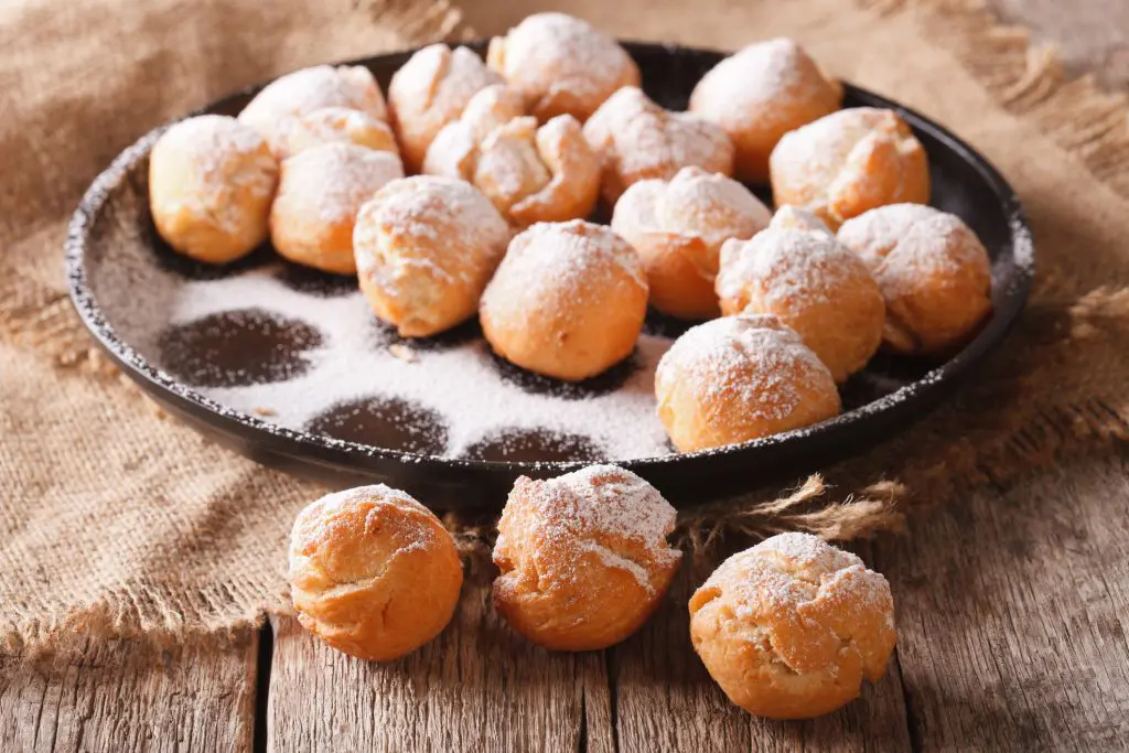 Plate of Italian castagnole—small round fried pastries dusted with powdered sugar—on a rustic wooden surface with burlap cloth.