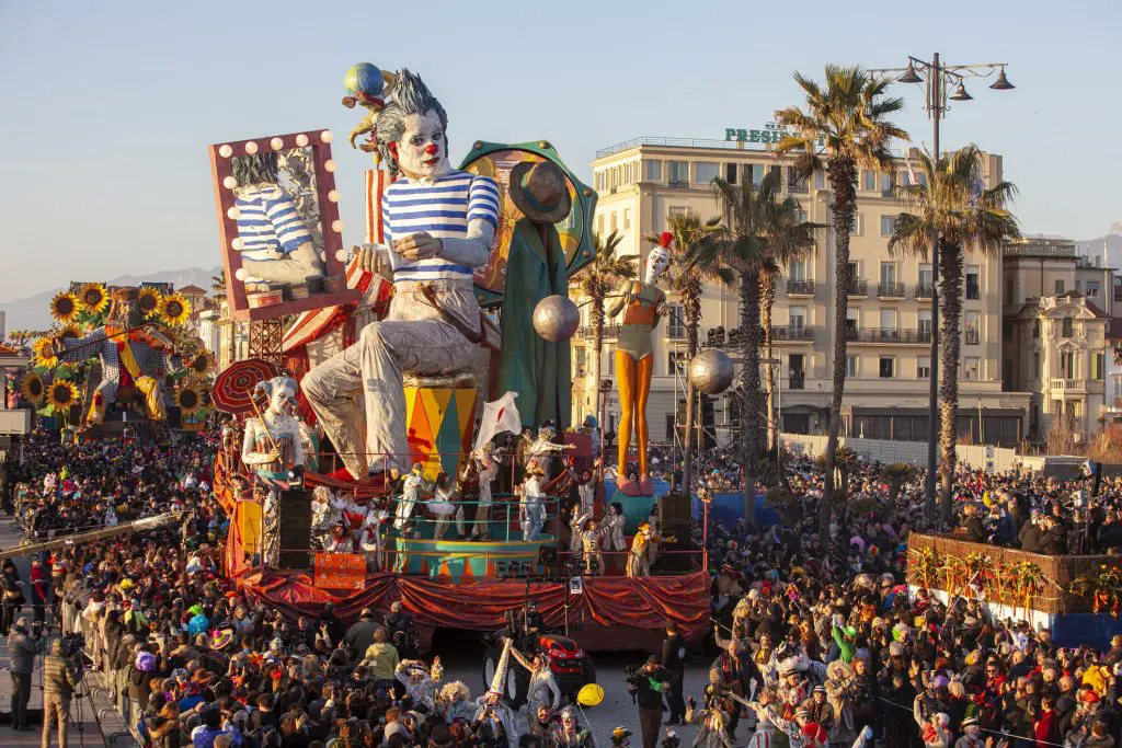 “Colorful parade at the Viareggio Carnival in Italy, featuring giant papier-mâché clown floats, performers in costume, and large crowds gathered along the palm-lined street.”