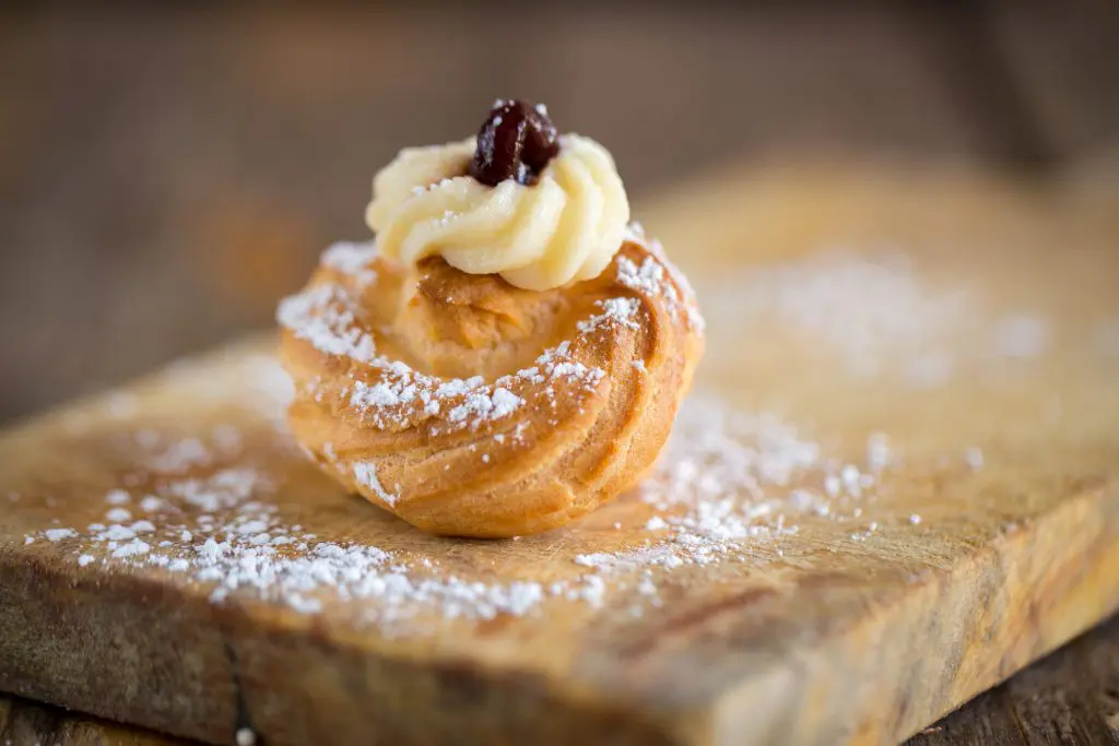 “Close-up of an Italian zeppola pastry topped with custard and a cherry, dusted with powdered sugar and served on a wooden board.”