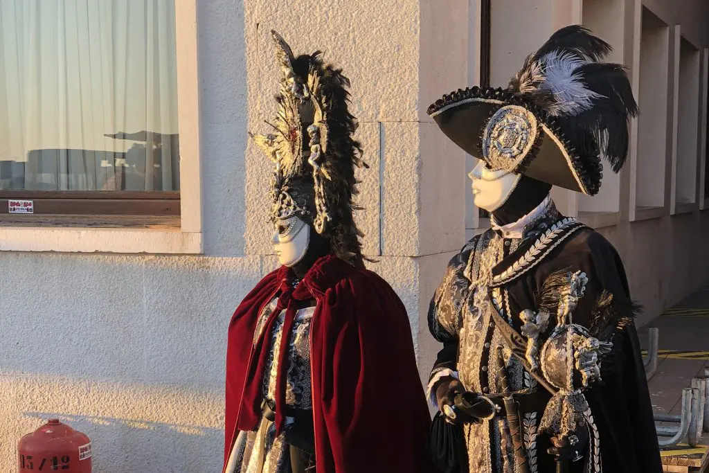 Two people in elaborate Venetian carnival costumes with ornate masks, feathers, and baroque-style embroidery, standing against a light-colored building wall during sunset