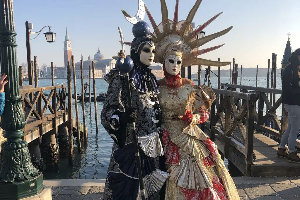 Two people in elaborate Venetian carnival costumes with ornate masks, feathers, and baroque-style embroidery, standing against a light-colored building wall during sunset