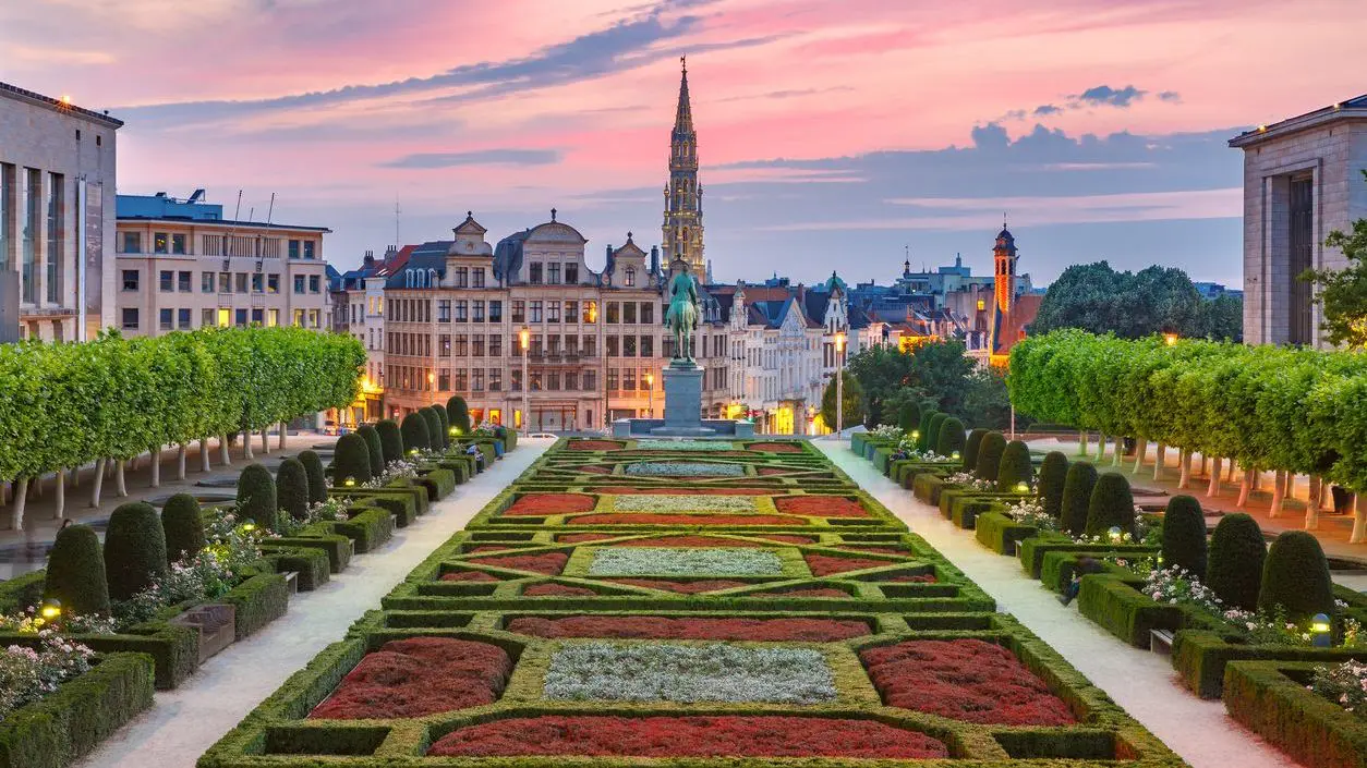 The Old Town of Brussels under a lilac sunset sky with a manicured garden in the foreground.
