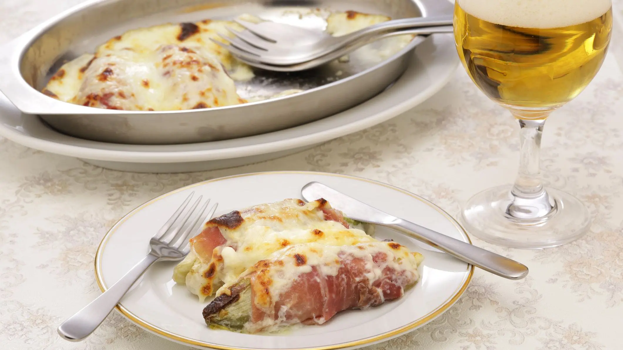 A white dish with two endives wrapped in ham and topped with melted cheese. The baking pan with the rest of the baked food is in the background.