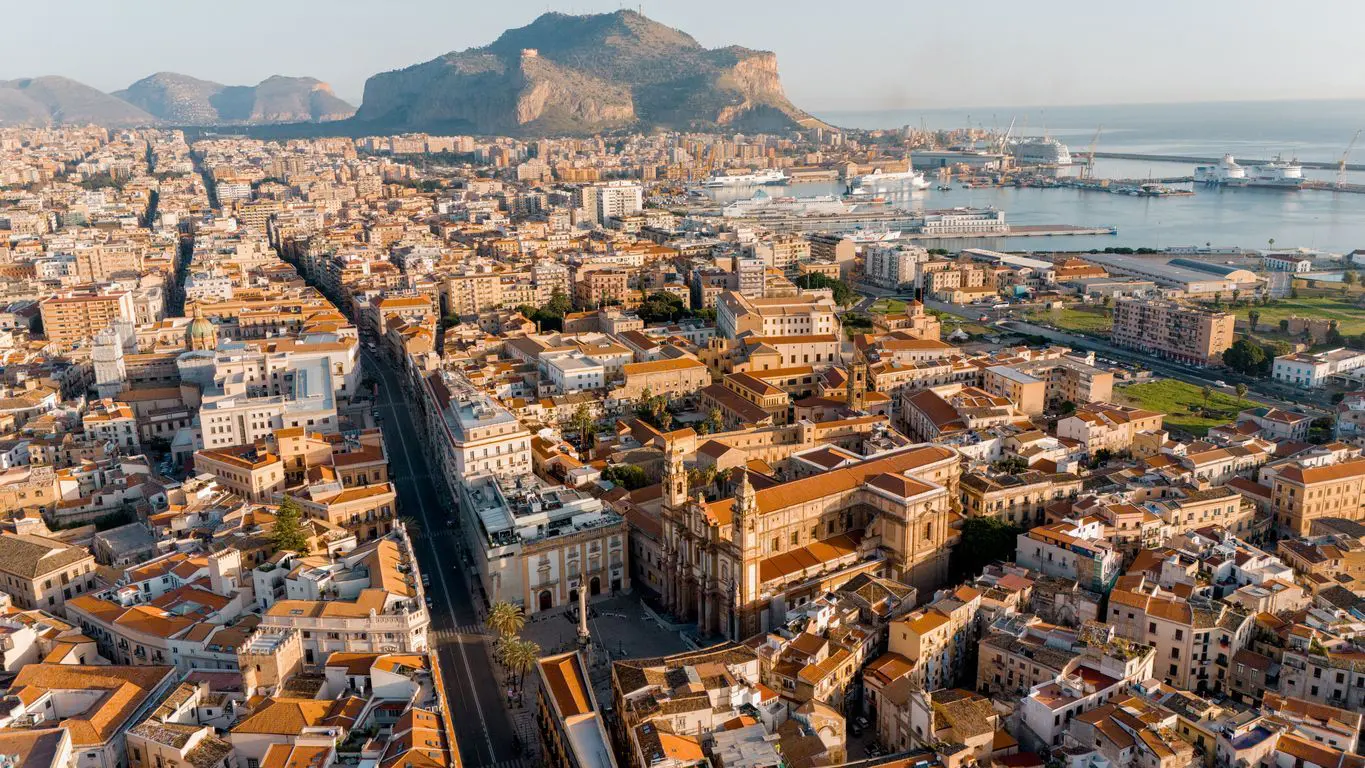An aerial panorama of Palermo, Sicily, showing the majestic Palermo Cathedral, Via Vittorio Emanuele, and the port stretching toward Monte Pellegrino in the morning light.
