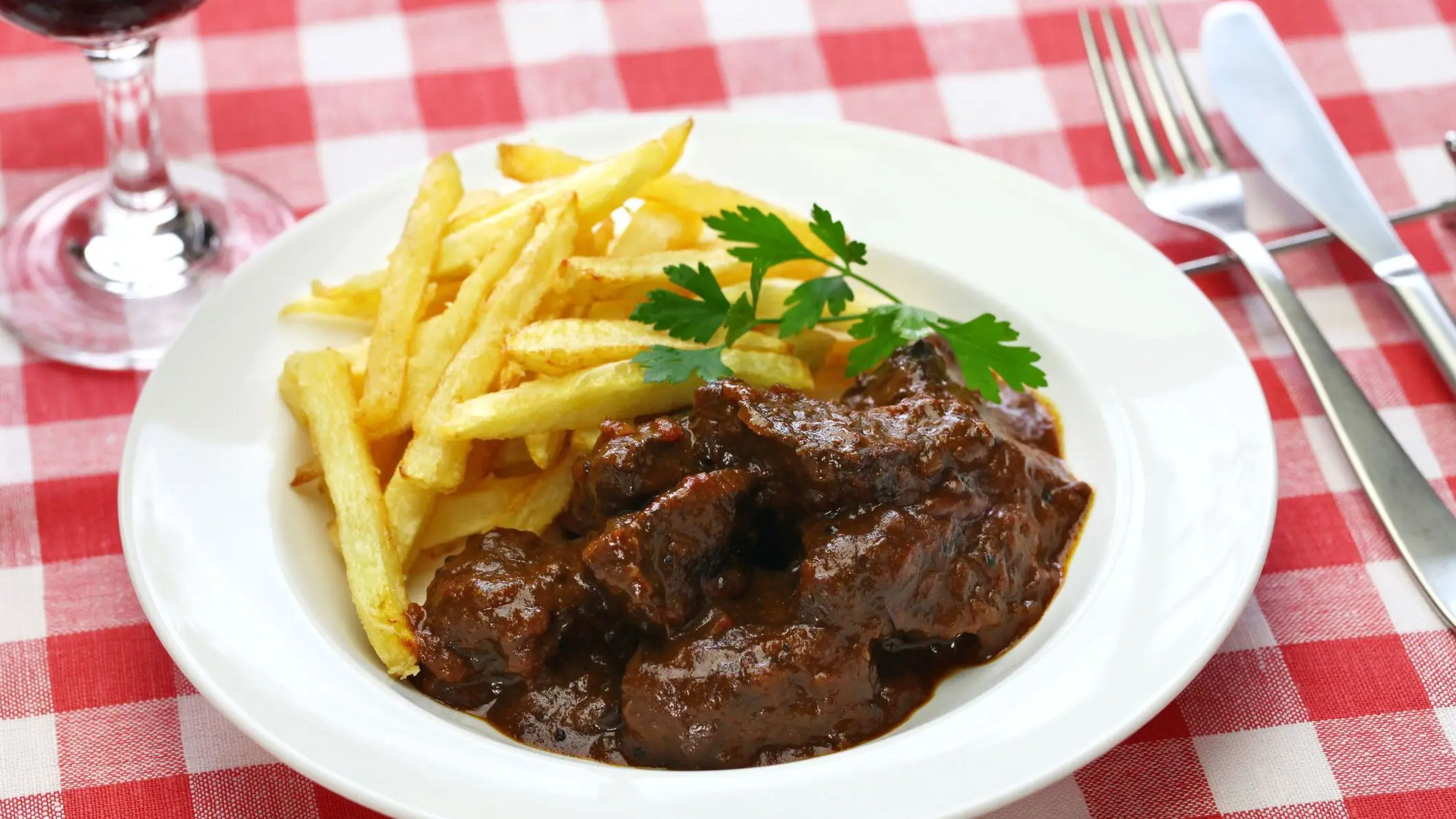 A white dish filled with beef and fries on a red-and-white checkered tablecloth.