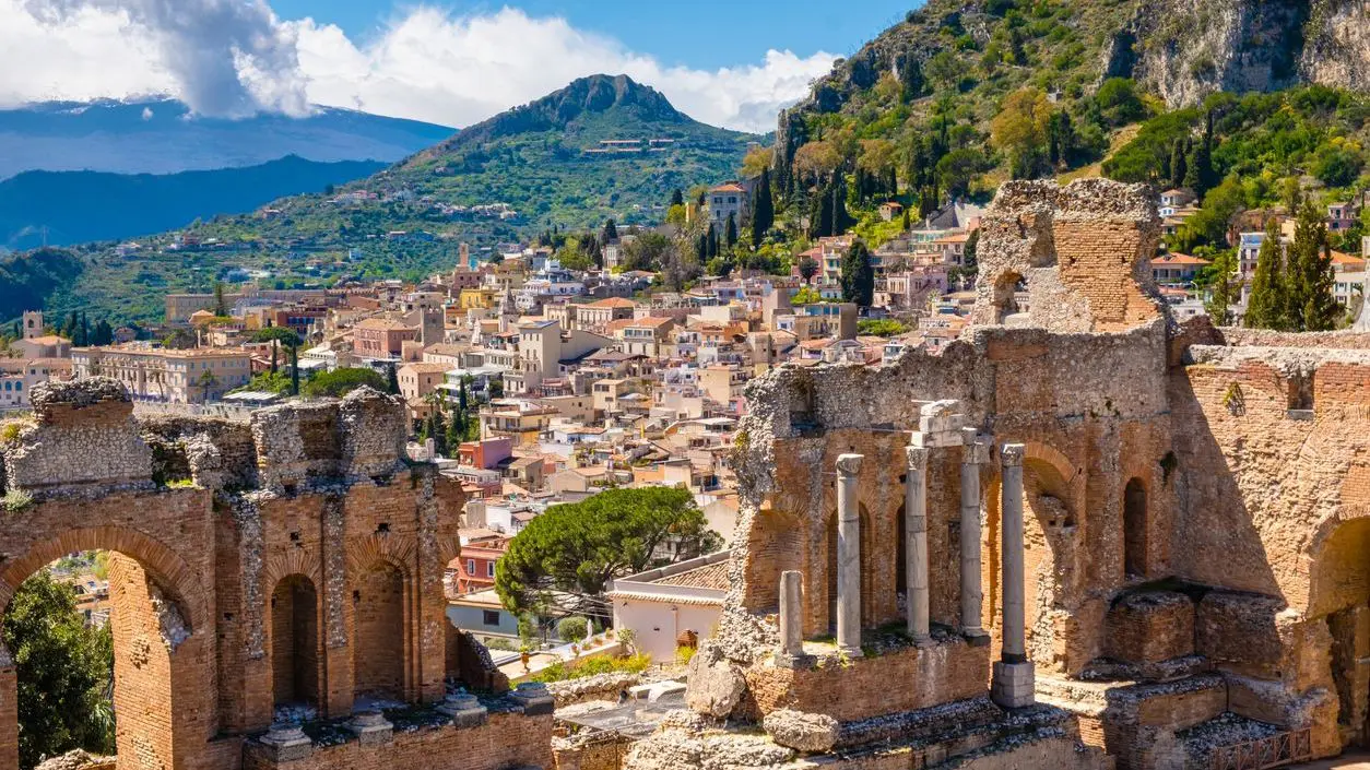 The ruins of an Ancient Greek theater, with the Old Town and Mount Etna covered with clouds in the background.