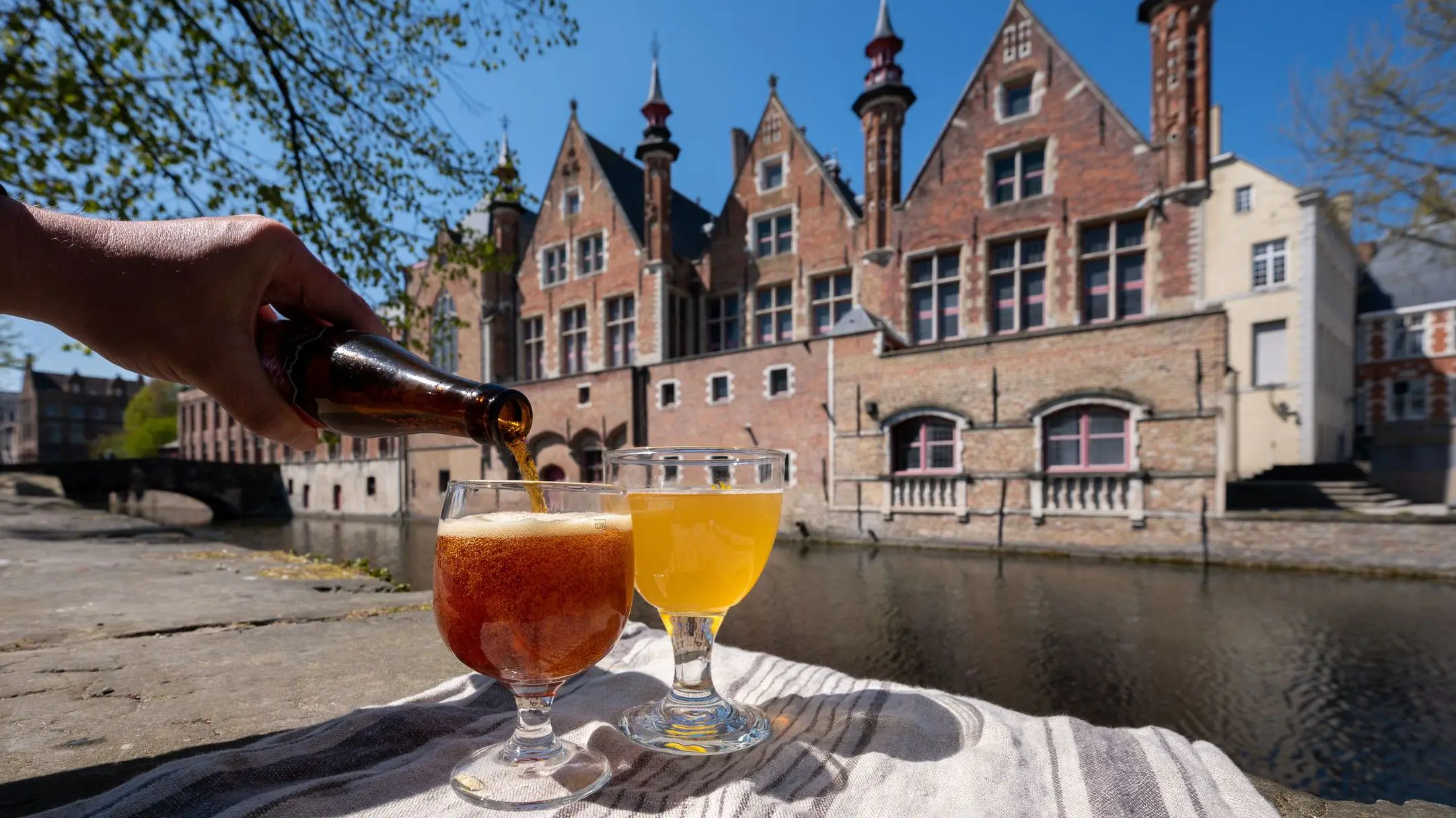 A hand serving beer in a glass next to another glass of beer with medieval buildings and a river in the background.