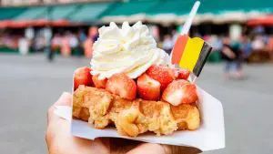A close-up of a Belgian waffle topped with strawberries, whipped cream, and a tiny paper flag of Belgium.