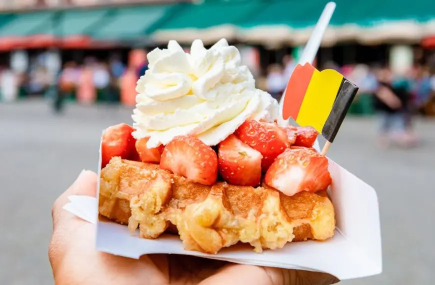 A close-up of a Belgian waffle topped with strawberries, whipped cream, and a tiny paper flag of Belgium.