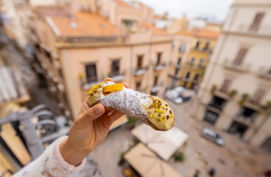 A hand holds a traditional Sicilian cannolo over a balcony with a view of old Palermo rooftops.