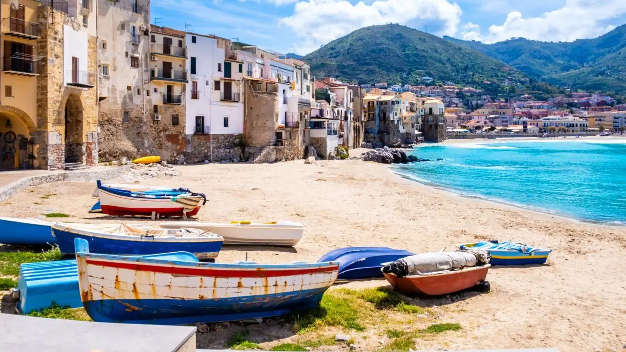 Fishing boats on a beach in Cefalu, a medieval town on Sicily Island, Italy. There are historic buildings on the edge of the sand, clear turquoise sea water and mountains.