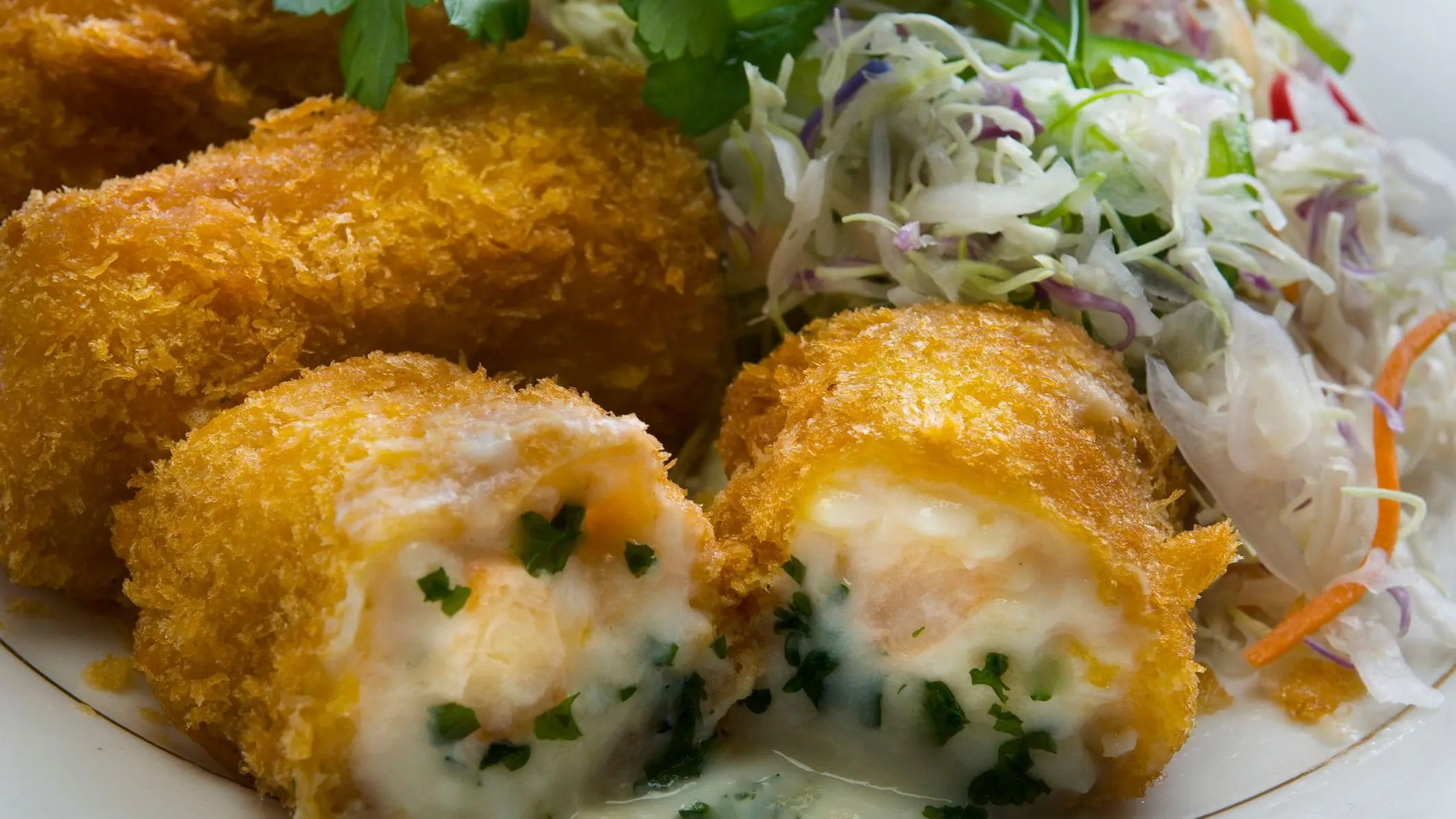 A close-up of a golden croquette cut in half. There's white sauce, shrimp, and herbs inside.