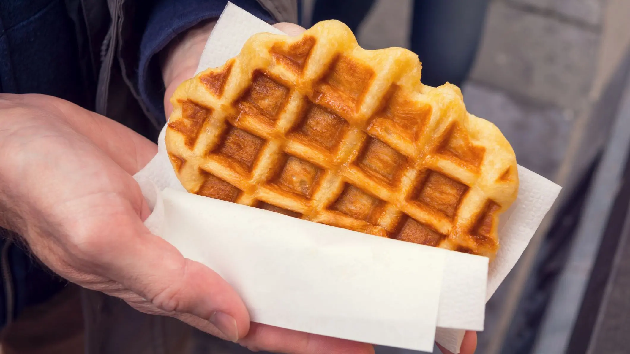 A man's hands are holding a fresh and warm Liege-style Belgian waffle wrapped in white paper.