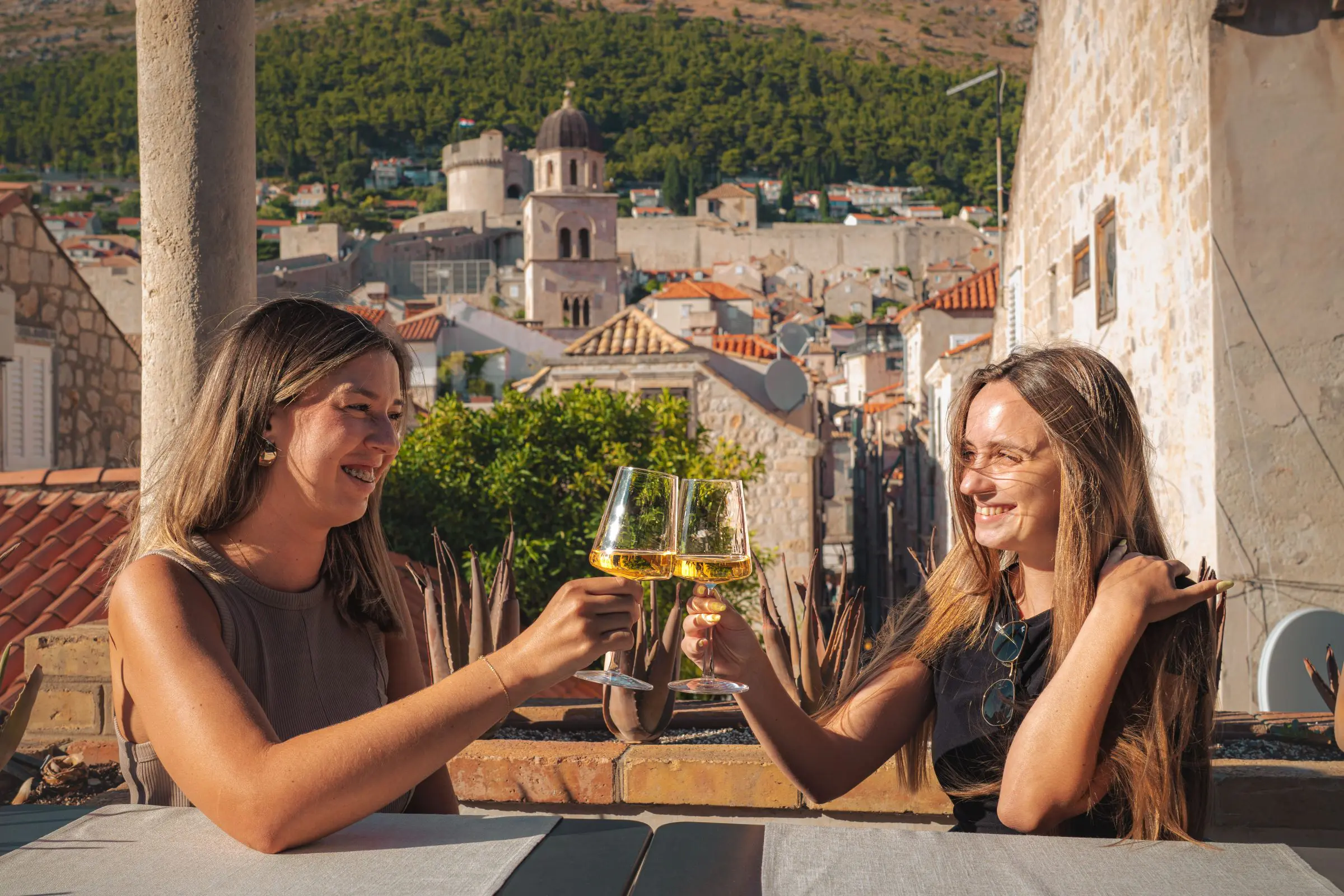 Two young women sitting at an outdoor terrace with a view on a sunny day, clinking their white wine glasses and smiling.