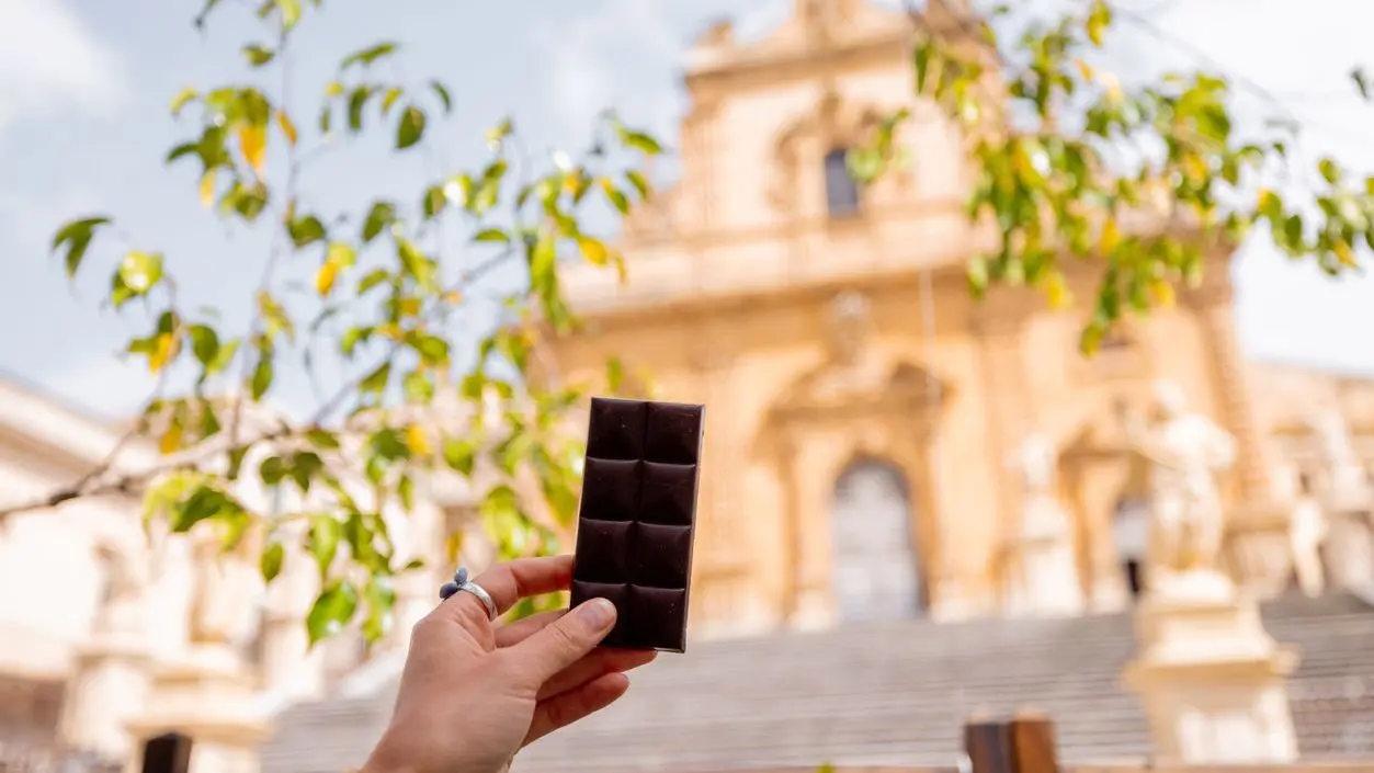 A close-up of a hand holding a traditional Modica chocolate bar with a blurred view of Saint Peter Cathedral in the background.
