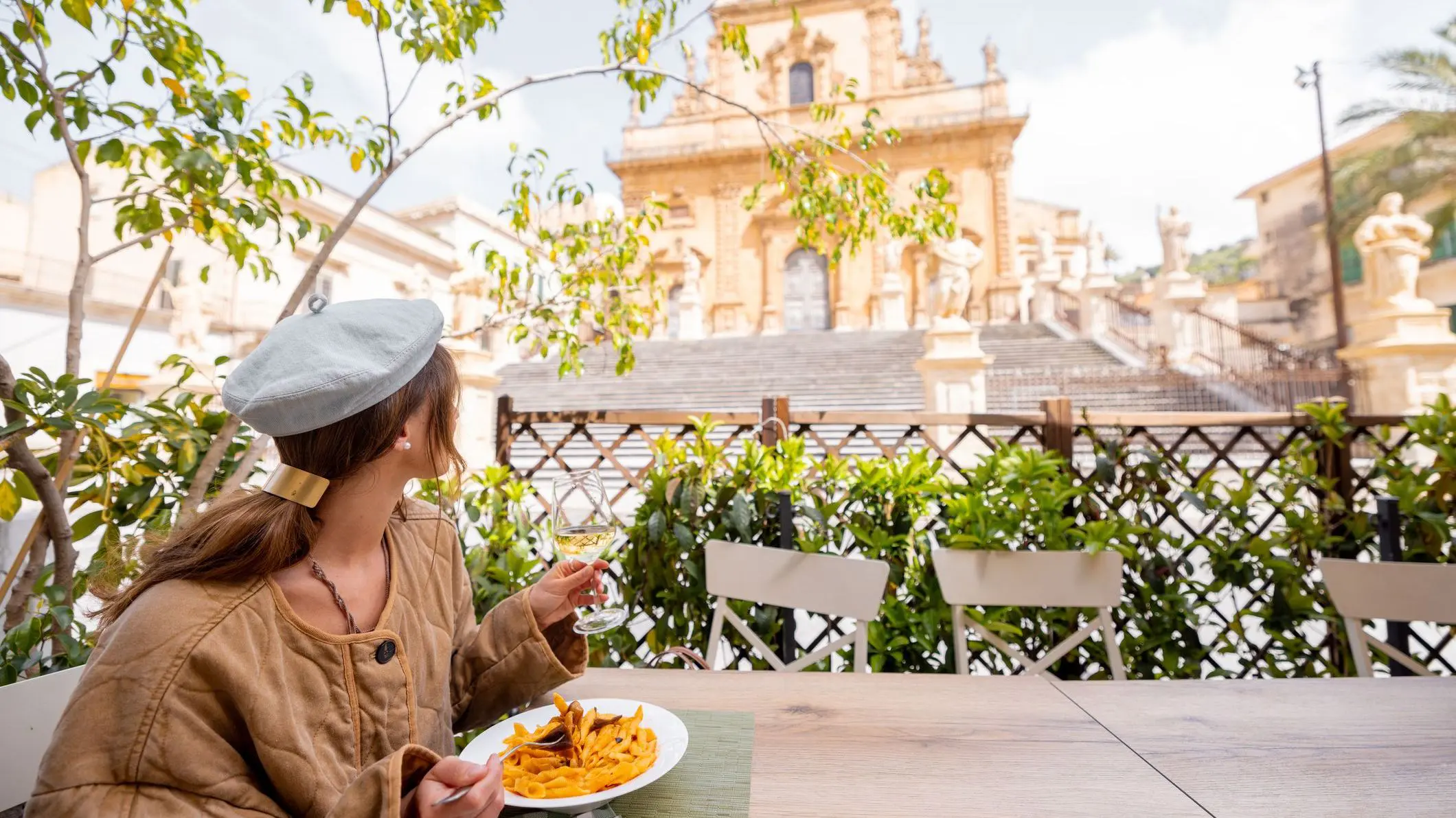 A woman enjoys a plate of traditional Sicilian pasta alla Norma and a glass of white wine at an outdoor café in Modica, with Saint Peter Cathedral in the background.