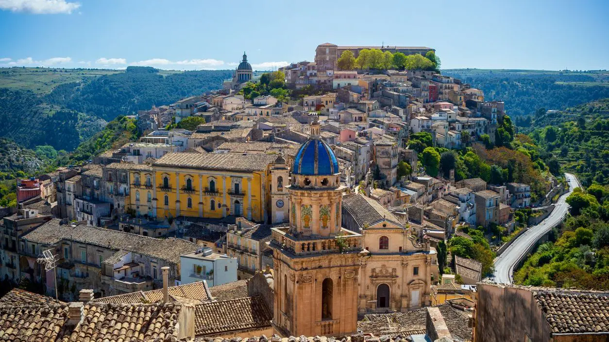 A panoramic view of the historic center of Ragusa, Sicily, Italy, with the tower of the blue-domed Santa Maria dell'Itria church, a UNESCO World Heritage Site, in the foreground.