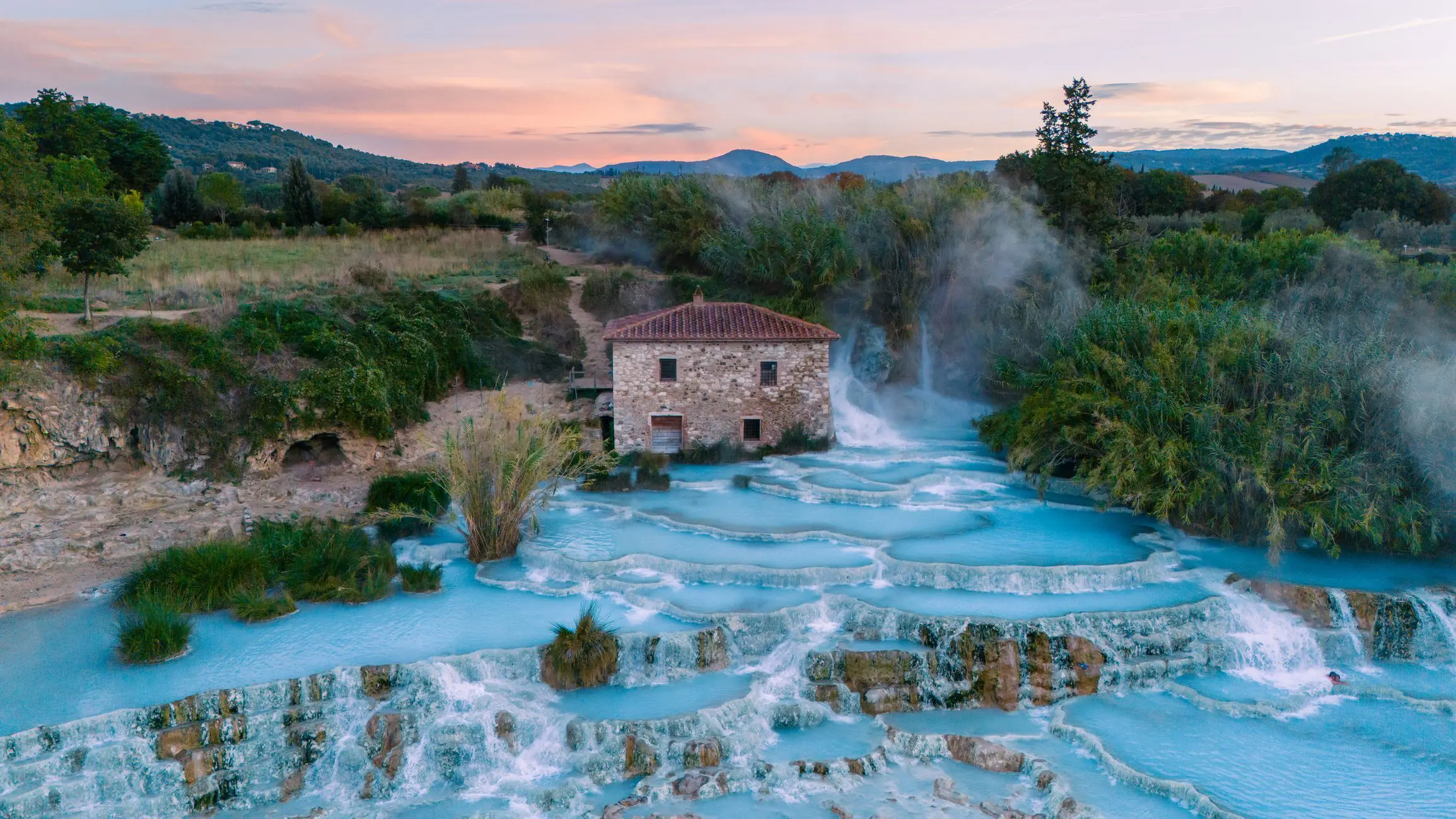 The Saturnia thermal baths surrounded by lush greenery and a charming stone house. The sun sets, casting warm hues over this tranquil Tuscan landscape.
