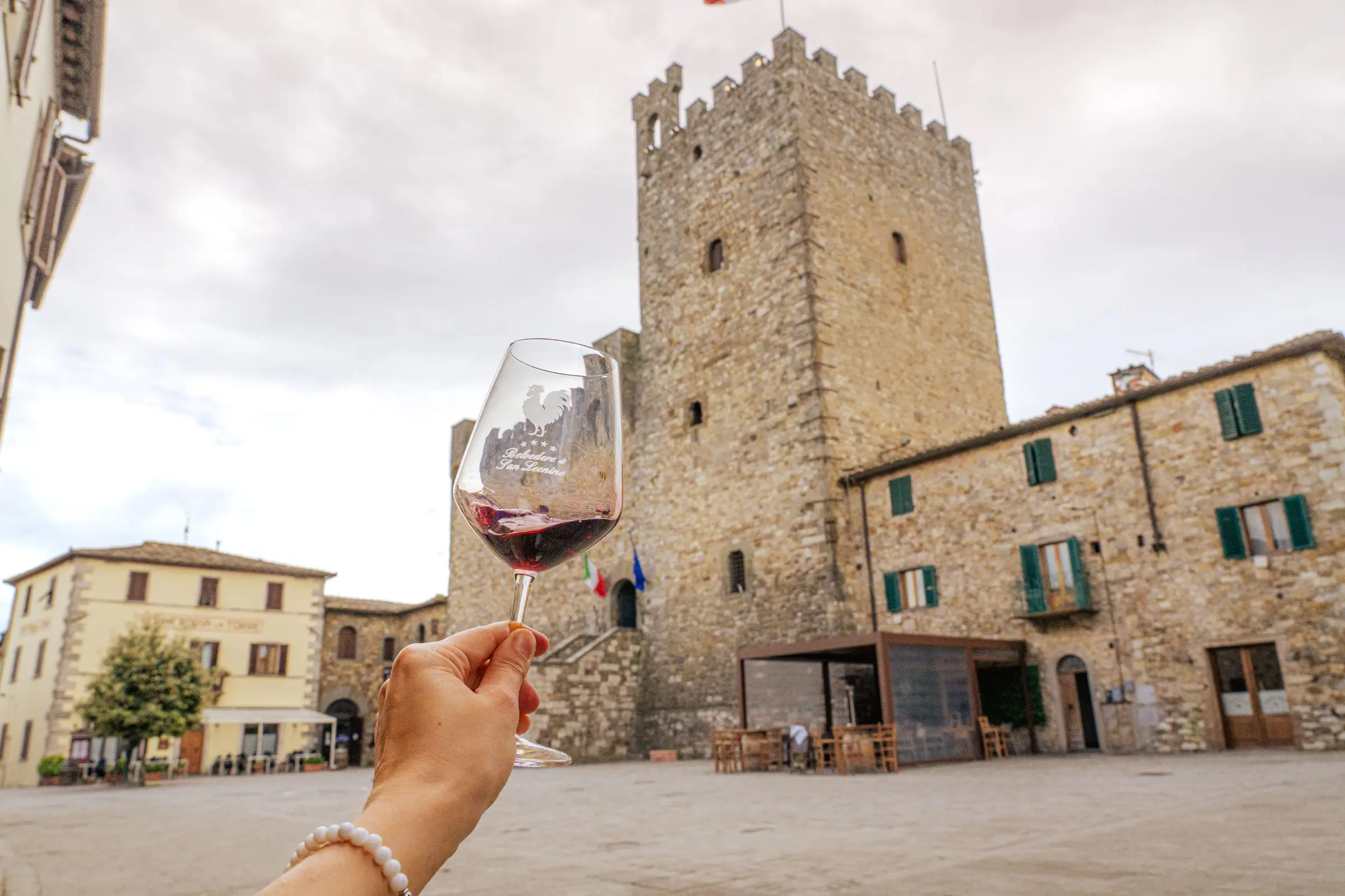 A hand is holding a glass of Chianti wine in front of a traditional Tuscan building.